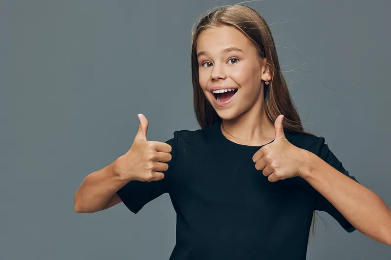 Teenage girl smiling and showing thumbs up with both hands in casual black t shirt on gray background for happiness and success expression.