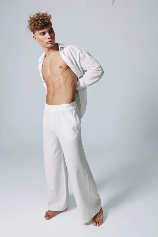 Young man with curly hair standing barefoot wearing white pants and open shirt exposing toned muscular torso against plain light background for fashion and fitness concept.