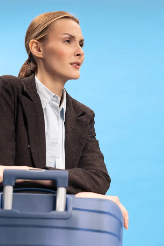 Young woman with suitcase looks thoughtfully to the side against blue background. Concept of travel, planning, and journey with modern luggage and casual outfit in studio lighting.