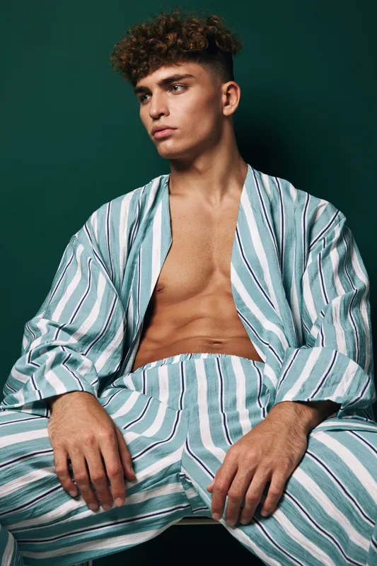 Young man with curly hair wearing striped robe and trousers sitting against dark background, neckline open with exposed chest, casual relaxed pose showing concentrated facial expression.