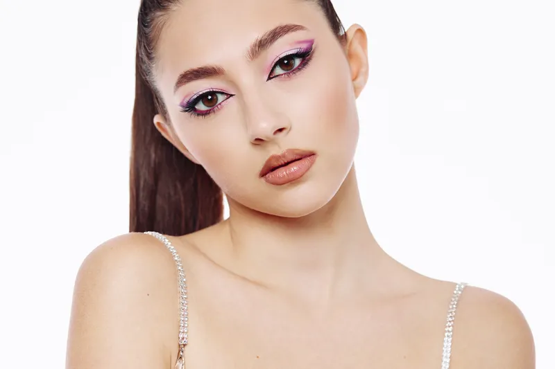 Close-up portrait of a young woman with creative pink eyeshadow and nude lips wearing a rhinestone strap top on a white background. Concept of beauty fashion and makeup artistry.