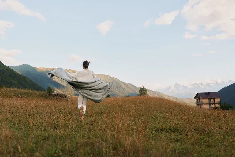 A young woman in white clothes runs joyfully through a grassy field with a blanket spread behind her under a bright sky in a mountainous landscape.