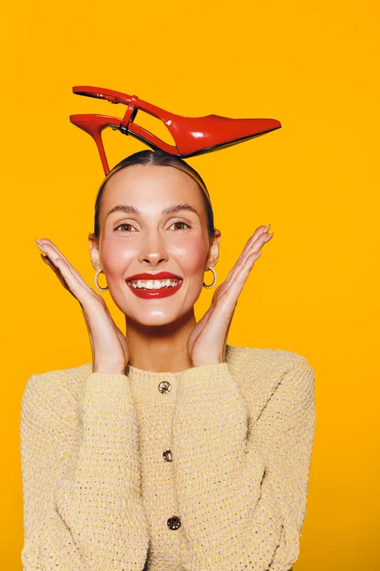 Smiling woman with red lipstick and hoop earrings holds red high heel shoe on her head against bright yellow background. Concept of fashion, style, and playful mood.