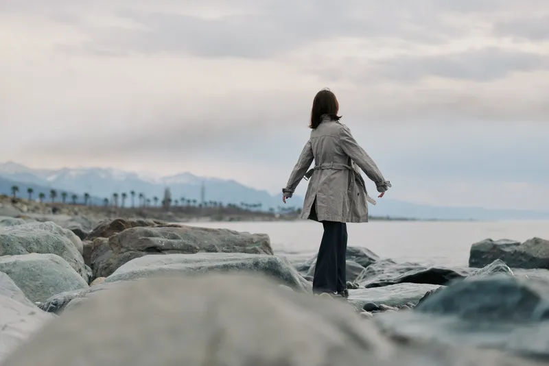 Woman in a gray coat walks on rocky beach near calm sea under cloudy sky. Mountains and palm trees are visible in the distance. Concept for travel, nature, and solitude.