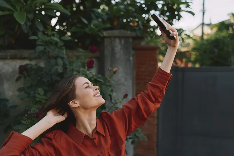 Woman enjoying lifestyle moments outdoors, taking a selfie with smartphone, wearing a red shirt, smiling in a relaxed pose among greenery, bright natural light, candid portrait of everyday joy