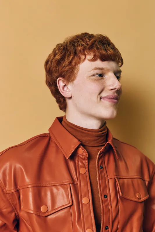 Young man with red hair wearing a brown leather jacket and turtleneck sweater posing looking sideways against neutral background. Portrait of confident smiling male model indoor.