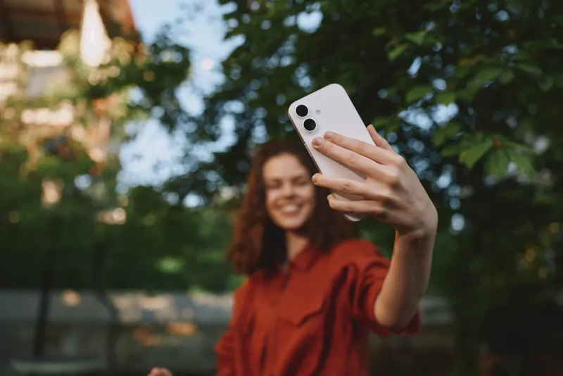 Smiling woman outdoors taking a selfie in a casual red shirt, surrounded by greenery. Emphasizes lifestyle, wellness, and moment of connection. Soft daylight highlights facial features and creates a