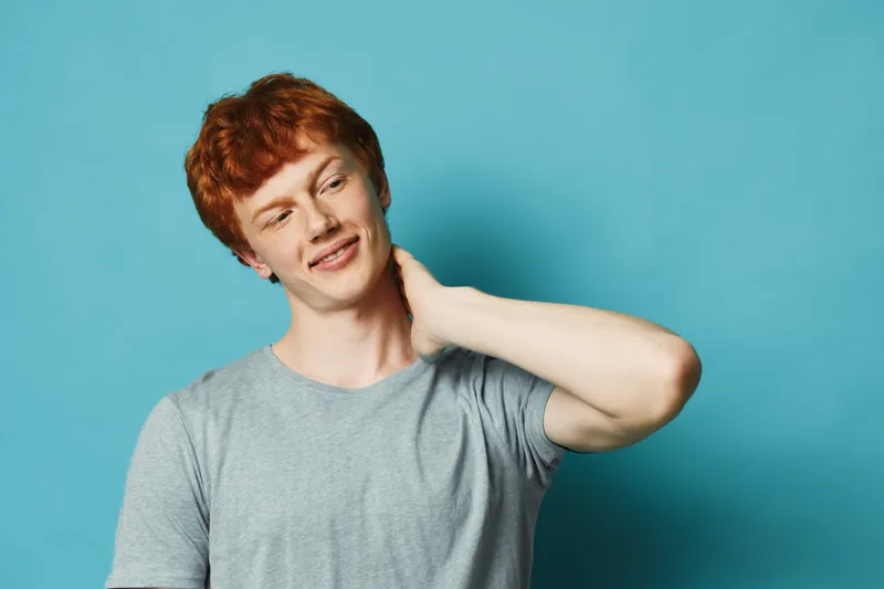 Young man with red hair wearing gray t shirt touching neck with hand feeling relaxed and thoughtful on blue background portrait of casual male with smile and comfortable pose.