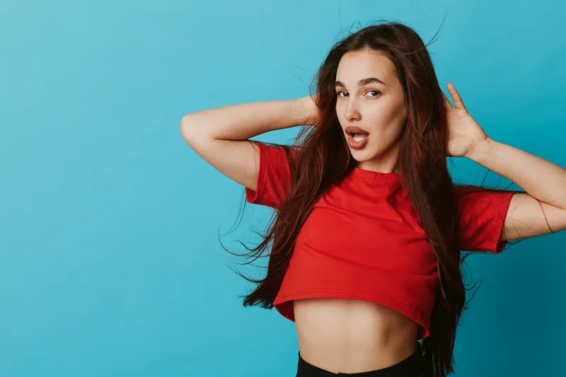 Young woman posing with hands behind head and open mouth expression against blue background. Casual style with red cropped t shirt and long dark hair for fashion portrait or lifestyle.