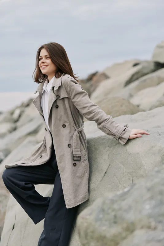 Smiling woman in trench coat and dark pants sits on large rocks by the sea. Overcast sky and natural scenery create a calm outdoor atmosphere with casual fashion concept.