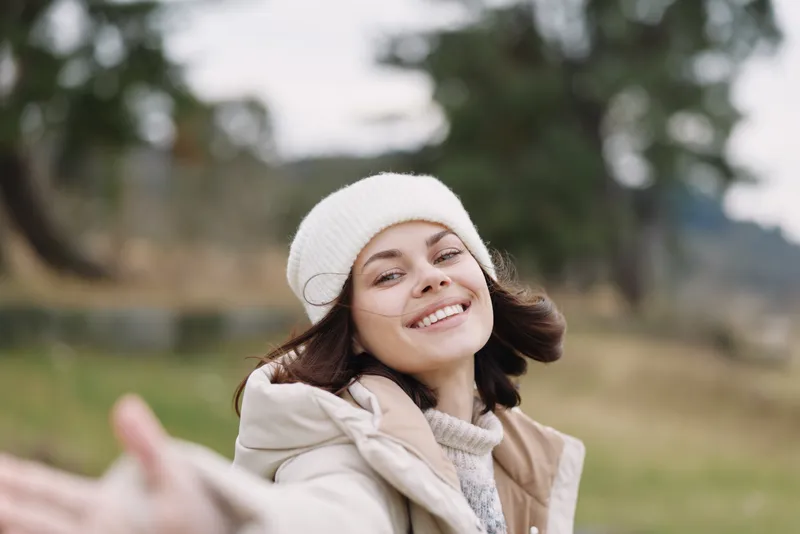 Portrait of a woman outdoors, smiling, reaching, in a beanie and casual jacket, captured in a sunny autumn park with gentle breeze