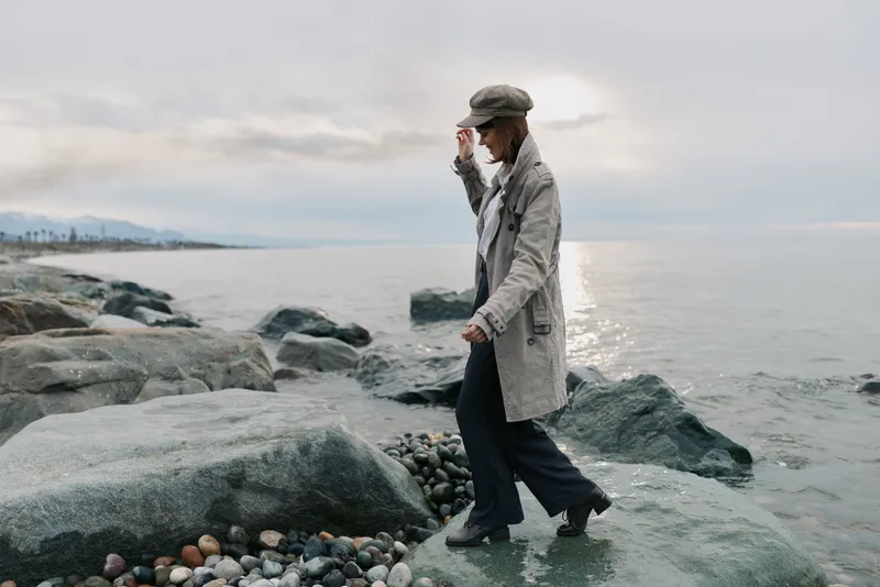 Woman in a grey coat and hat walks on rocks by the sea during cloudy day. Calm coastal landscape with pebbles and water in the background. Concept for autumn or outdoor lifestyle.