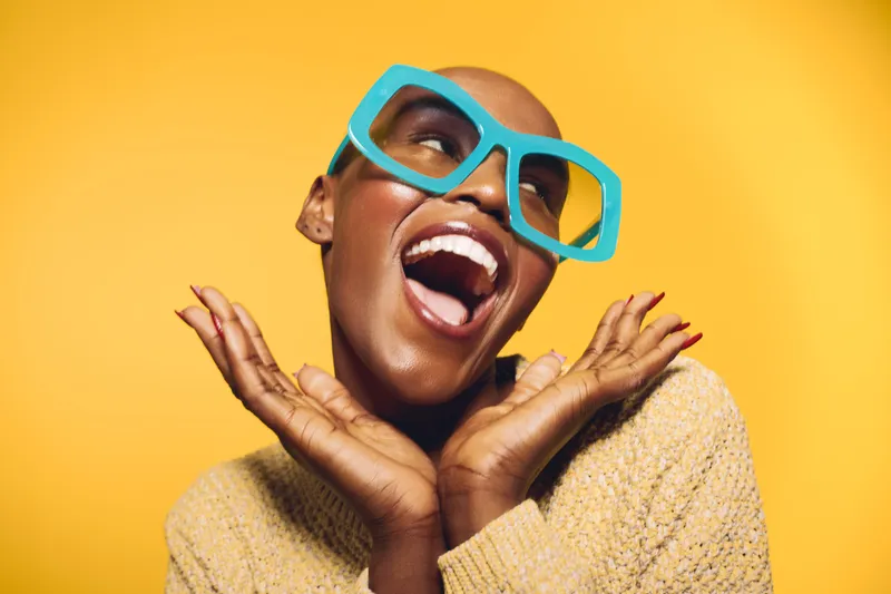 Happy dark skinned African woman with bald head and bright blue glasses laughing joyfully on yellow background. Concept of positive emotions, confidence, beauty, and fashion accessories.