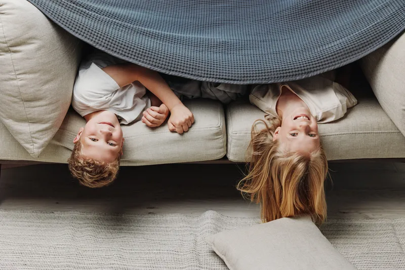 Two happy children lying upside down on a couch under a gray blanket, smiling and holding hands in a cozy living room setting.