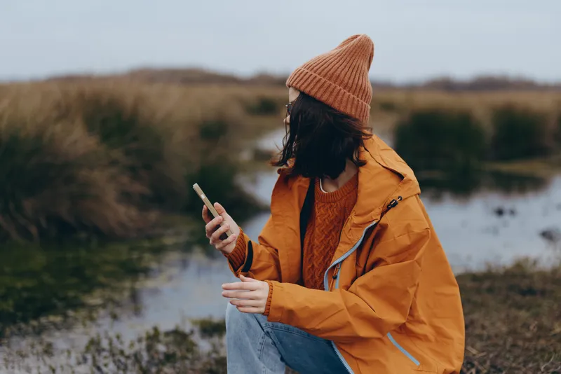 Woman outdoors countryside, orange jacket knit hat, holding cigarette, winter lifestyle moment, calm nature background, phone in hand, soft daylight, candid scene, fresh air feel