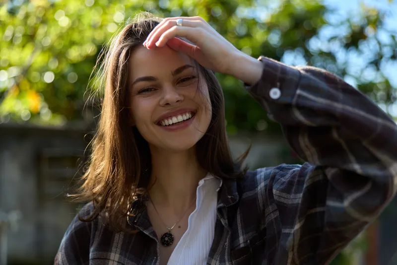 A cheerful woman in a plaid shirt outdoors, sunlit garden scene, lifestyle portrait showing casual confidence, warmth, and everyday happiness, relaxed mood