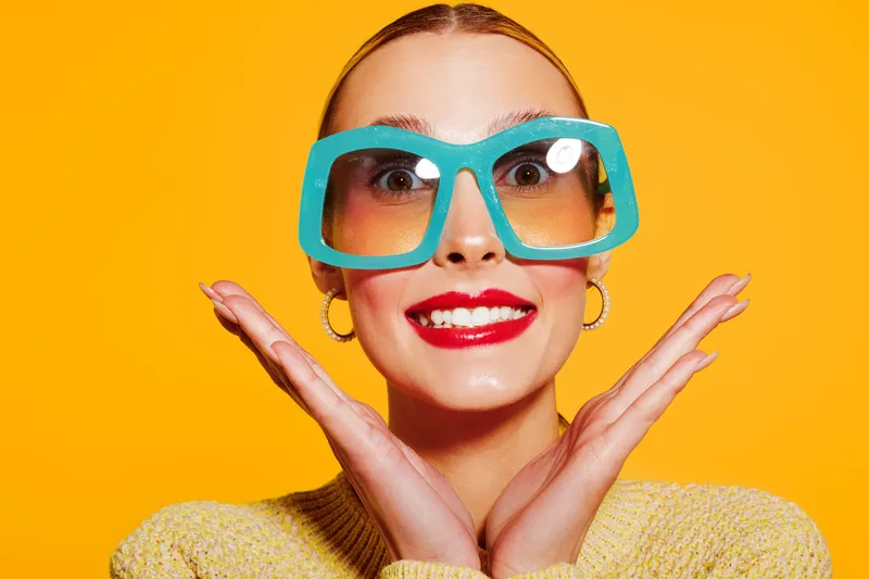 Smiling woman with large blue sunglasses and red lipstick poses with hands near face on bright yellow background. Concept of fashion, beauty, and summer style portrait.