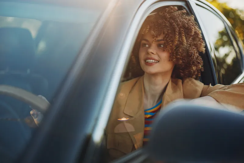A young woman with curly hair sits in a car, smiling as warm sunlight touches her beige jacket, creating a relaxed, cinematic, retro mood that hints at a confident, everyday moment.