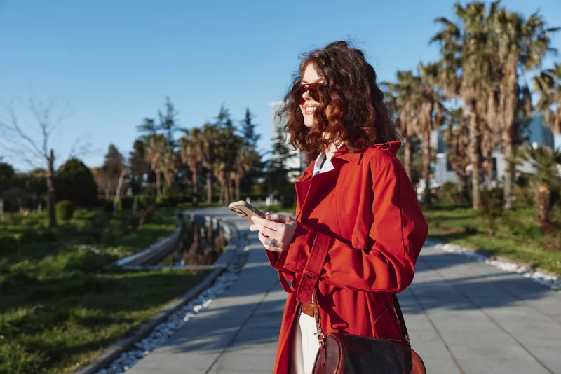 Young woman with curly brown hair wearing a red coat walks outdoors on a sunny day while holding a smartphone and smiling confidently.