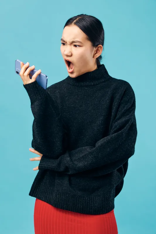 Woman in black sweater holding smartphone with angry expression shouting at device on blue background for communication or conflict in technology and emotions.