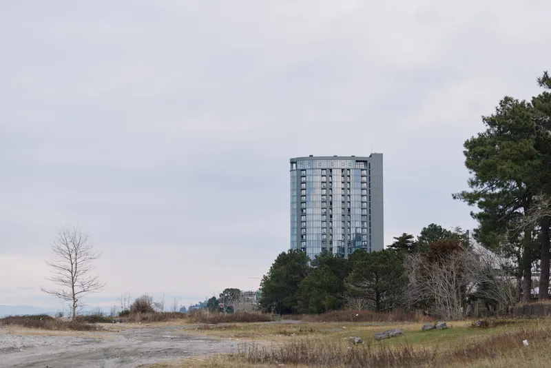 Tall high rise building rises over a flat field with sparse trees under a cloudy sky, creating strong urban contrast with the natural landscape and horizon.