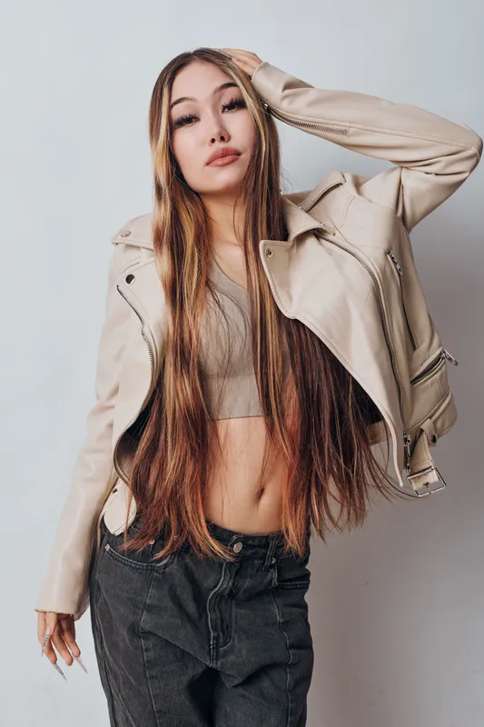 Young woman with long hair wearing cropped top and beige leather jacket posing indoors. Fashion model with casual style in black jeans and neutral background for portrait photography.