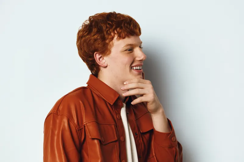 Young man with red hair wearing brown leather jacket smiling and looking sideways with hand near chin against white background for casual portrait and fashion style.