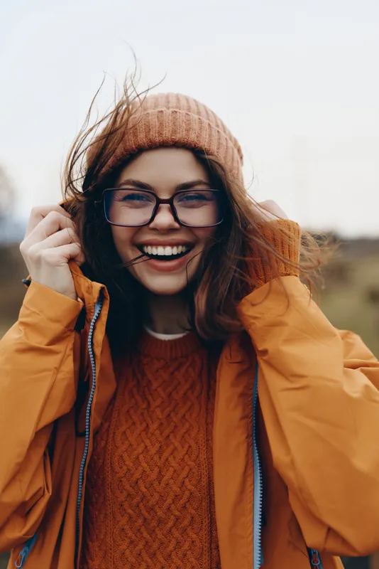 Smiling woman lifestyle portrait outdoors in autumn, orange knit hat and glasses, cozy coat, hands near ears, warm light, close up, casual winter style, happy
