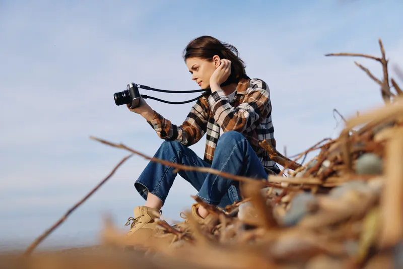 Photography, camera, outdoor, woman sits among driftwood by the sea, focused on landscape with calm mood, casual plaid outfit, natural light and reflective moment.