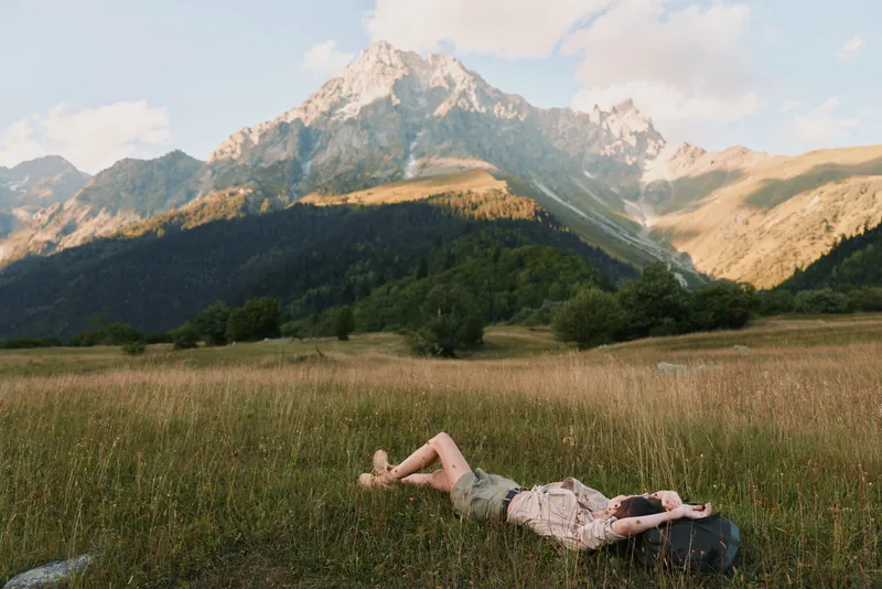 Young woman lies in a sunlit meadow with mountains in the background, relaxed pose and peaceful expression amid tall grass, a gentle breeze, warm light, natural colors, distant pines, and wide open