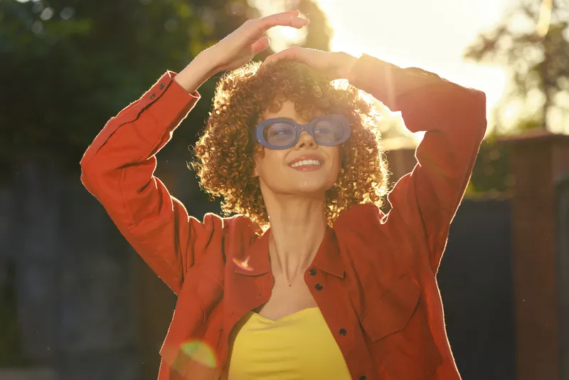 Young woman with curly hair in an orange jacket and yellow top shields her eyes with sunglasses under bright sun in a park, conveying carefree summer vitality