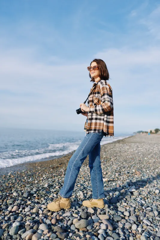 Woman walking along a pebbled beach in a casual plaid shirt, jeans and boots, holding a camera, under a sunny sky with calm sea and open horizon.