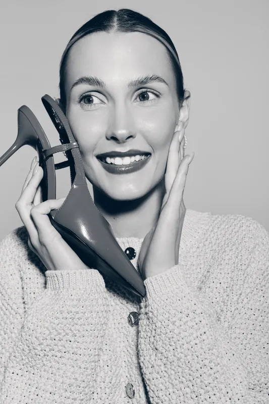 Young Caucasian woman with fair skin holding red high heel shoes near her face, smiling and posing in cozy knitted sweater. Concept of fashion, style, and beauty accessories.