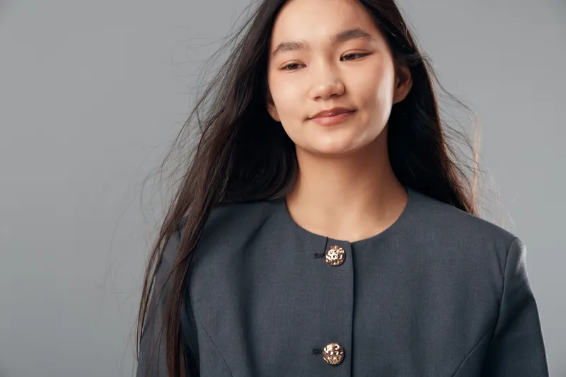 Young woman with long dark hair wearing gray jacket with decorative buttons looking down thoughtfully isolated on gray background, portrait and casual fashion style for lifestyle concept.