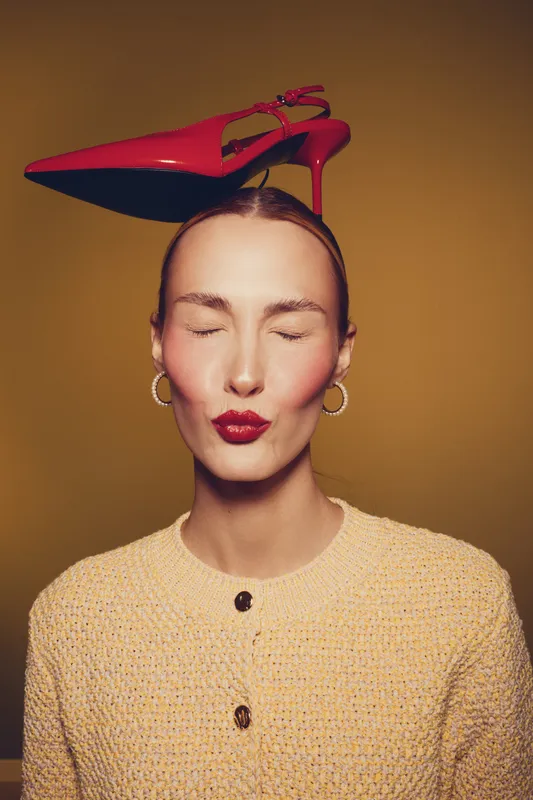 Young woman with closed eyes and red lipstick balances red high heel shoe on her head wearing yellow knitted sweater and gold hoop earrings on brown background.