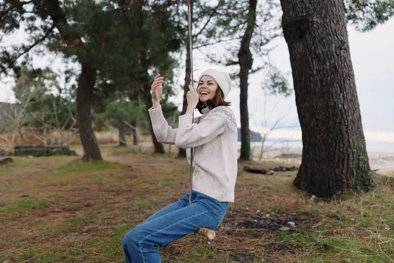 Young girl on a swing beside tall trees in a coastal forest, wearing a knit sweater and beanie, capturing a playful moment in nature.