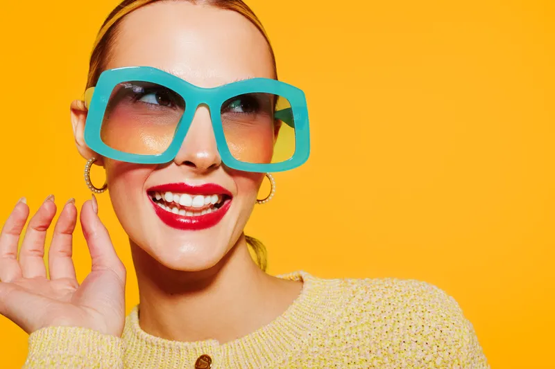 Smiling woman with large turquoise sunglasses and red lipstick poses against bright yellow background. Fashion concept with cheerful mood, colorful style, and trendy accessories in studio.
