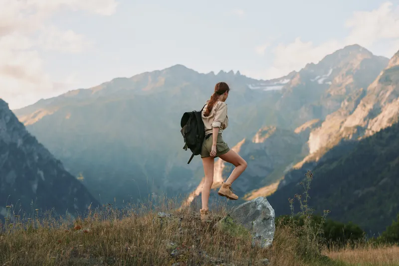 A woman hikes along a mountain trail with a backpack, stepping over a rock, framed by rugged peaks and wildflowers, capturing a moment of adventure and nature.