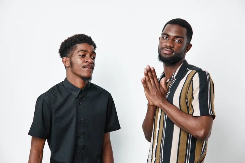 Two African American men with dark skin posing on white background, one wearing black shirt and the other striped shirt, showing casual style and confident expressions.