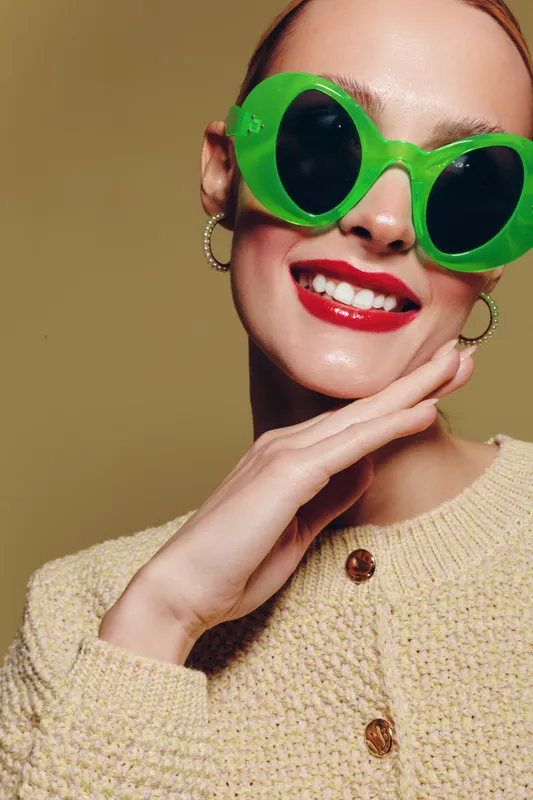 Smiling woman with bright red lipstick wearing large green sunglasses and beige knitted sweater poses with hand near face on neutral background. Concept of fashion, style, and confidence.