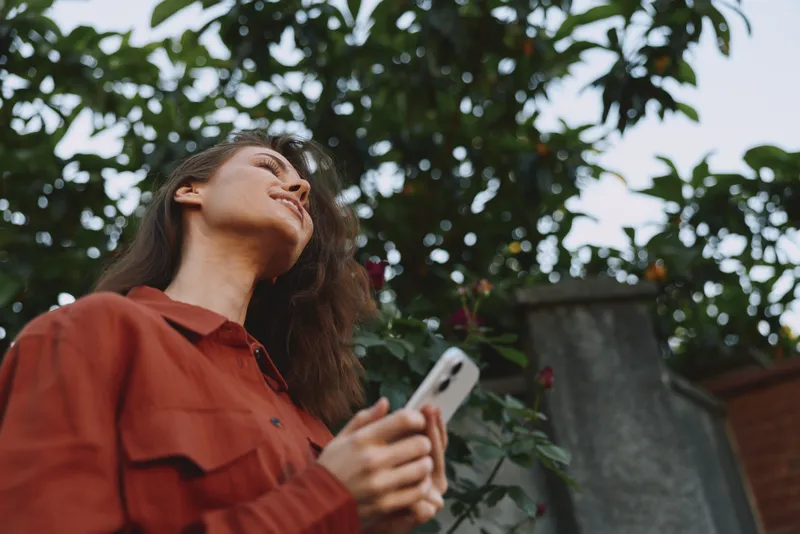 Woman in a casual red shirt gazes upward while holding a smartphone in an outdoor garden, capturing a relaxed lifestyle moment among greenery and warm sunlight