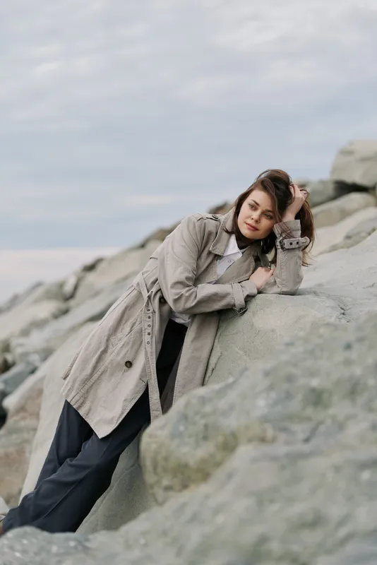 Young woman in knitted coat leans on rocks outdoors with closed eyes. Overcast sky creates calm atmosphere. Concept of autumn fashion, relaxation, and natural beauty.