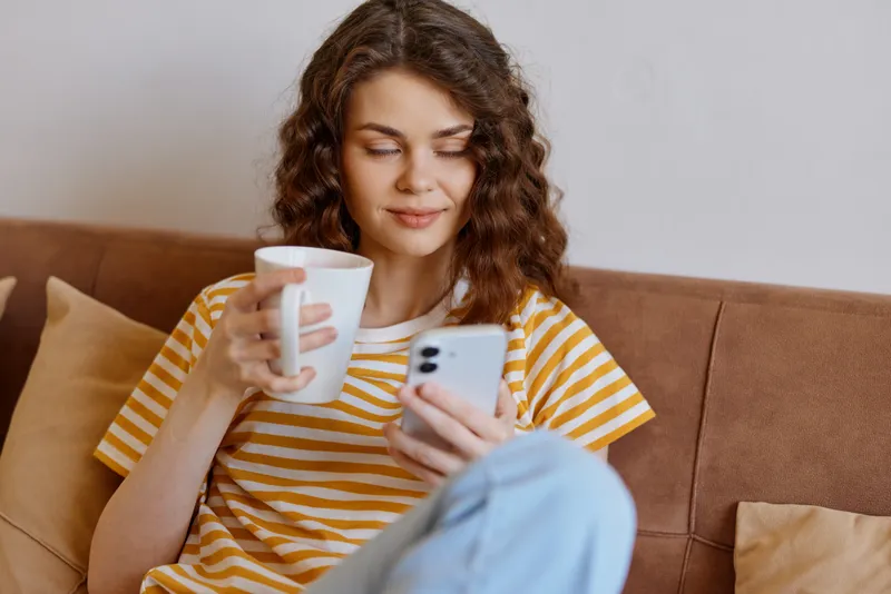 A woman relaxes at home on a couch, holding a mug and checking her smartphone, showing casual lifestyle and cozy comfort in a simple domestic scene.