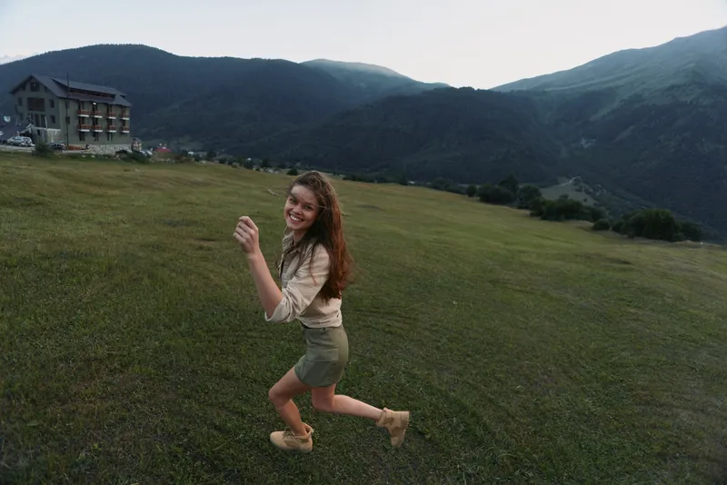 Young woman runs across an open meadow toward distant mountains, smiling as she enjoys a carefree outdoor moment in bright daylight.