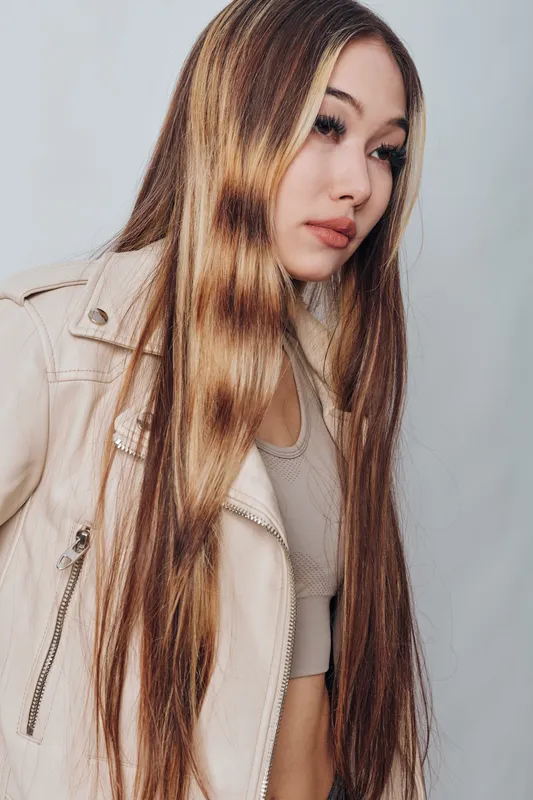 Young woman with long straight hair wearing beige jacket and crop top looking sideways with serious expression on plain background for fashion and beauty portrait.