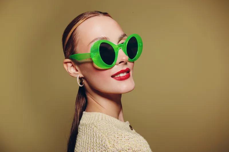 Stylish woman with green round sunglasses and red lipstick smiles confidently against beige background. Fashion portrait with trendy accessories and modern makeup look.