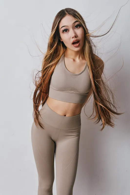 Young woman with long hair wearing beige sportswear posing with surprised expression on plain light background. Fitness, fashion and casual studio portrait for lifestyle concept.