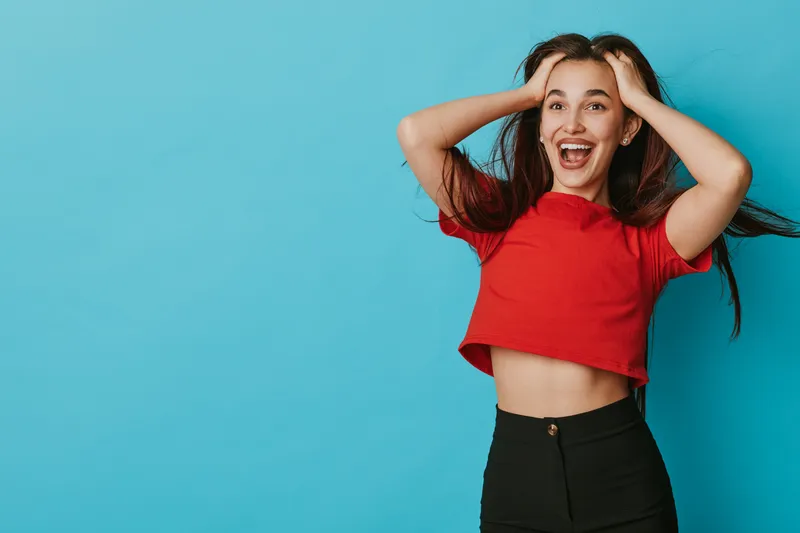 Young woman in red crop top and black pants with long hair smiling excitedly and holding head on blue background. Happiness, joy, and positive emotion in casual style portrait.