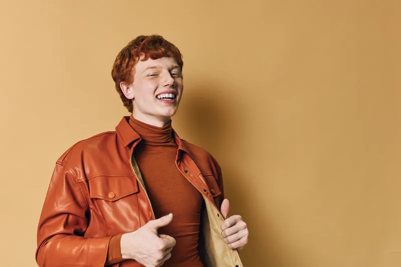 Young man with red hair and freckles smiling and laughing wearing a brown leather jacket and turtleneck in front of solid beige background for portrait and fashion.