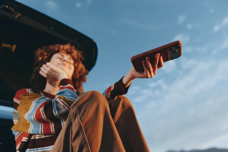 Woman lifestyle outdoors travel scene, curly hair woman in coat holds smartphone for selfie, arm raised toward blue sky, casual street travel moment, natural light, cinematic color
